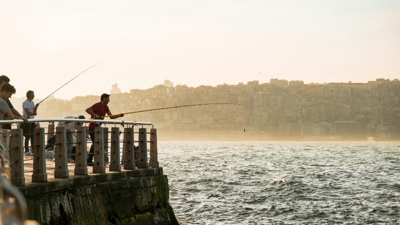 A group of men enjoy fishing from a pier by the sea with a distant cityscape backdrop during sunset., tags: driftfischen - pexel