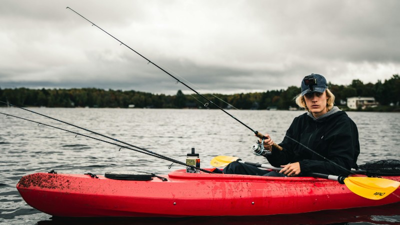 Young man fishing from a red kayak on a calm lake under cloudy skies., tags: drop-shot-rig - pexel